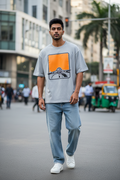 Man walking on a city street wearing a gray t-shirt with a graphic design, light blue jeans, and white sneakers.